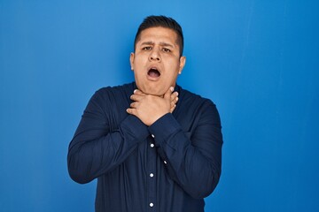 Hispanic young man standing over blue background shouting and suffocate because painful strangle....