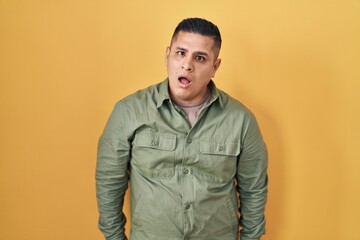 Hispanic young man standing over yellow background in shock face, looking skeptical and sarcastic, surprised with open mouth
