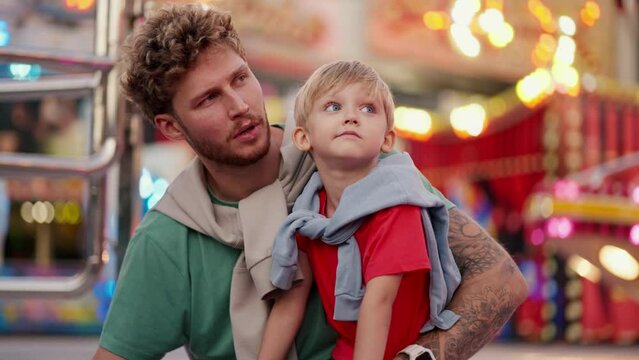 A little blond boy in a red T-shirt sits on his dad s leg and together they look at the bright and colorful attractions in the amusement park and communicate