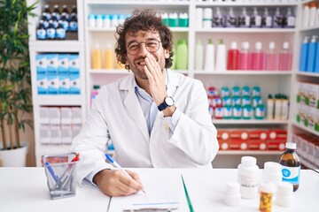 Hispanic young man working at pharmacy drugstore laughing and embarrassed giggle covering mouth...