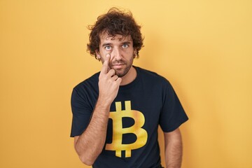 Hispanic young man wearing bitcoin t shirt pointing to the eye watching you gesture, suspicious expression