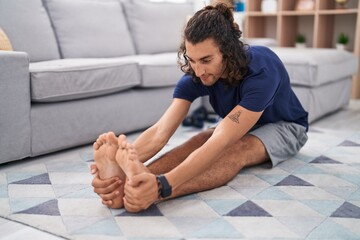 Young hispanic man stretching legs sitting on floor at home