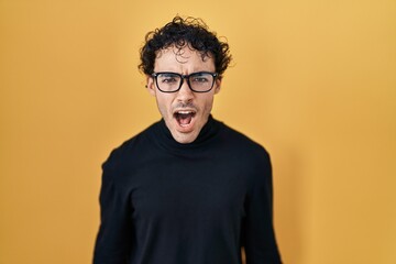 Obraz premium Hispanic man standing over yellow background angry and mad screaming frustrated and furious, shouting with anger. rage and aggressive concept.
