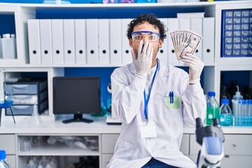 Hispanic man working at scientist laboratory holding money covering mouth with hand, shocked and afraid for mistake. surprised expression