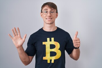 Caucasian blond man wearing bitcoin t shirt showing and pointing up with fingers number six while...
