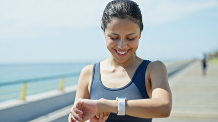 Young beautiful hispanic woman wearing sportswear looking smartwatch smiling at seaside - obrazy, fototapety, plakaty