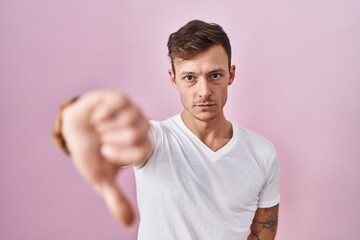 Caucasian man standing over pink background looking unhappy and angry showing rejection and negative with thumbs down gesture. bad expression.