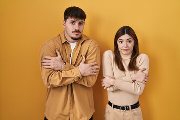 Young hispanic couple standing over yellow background shaking and freezing for winter cold with sad and shock expression on face