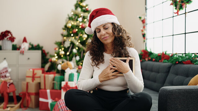 Middle age hispanic woman touching heart with photo sitting on sofa by christmas tree at home
