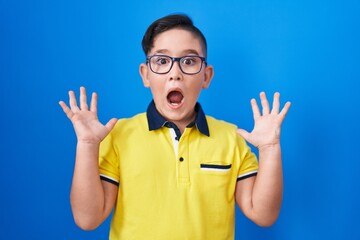Young hispanic kid standing over blue background celebrating victory with happy smile and winner expression with raised hands