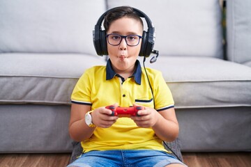 Young hispanic kid playing video game holding controller wearing headphones making fish face with...