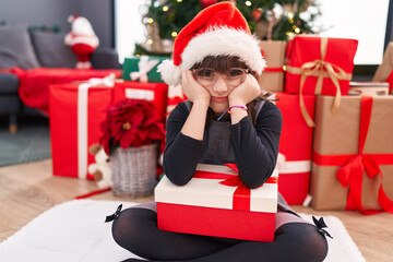 Adorable hispanic girl holding gift sitting on floor by christmas tree with unhappy expression at...