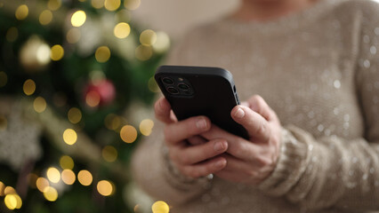 Middle age woman with grey hair using smartphone by christmas tree at home