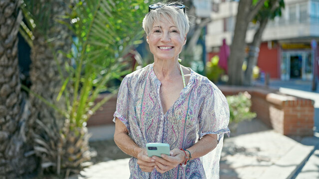 Middle age blonde woman tourist smiling confident using smartphone at street