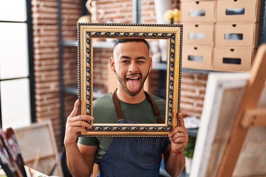 Young hispanic man sitting at art studio with empty frame sticking tongue out happy with funny expression.
