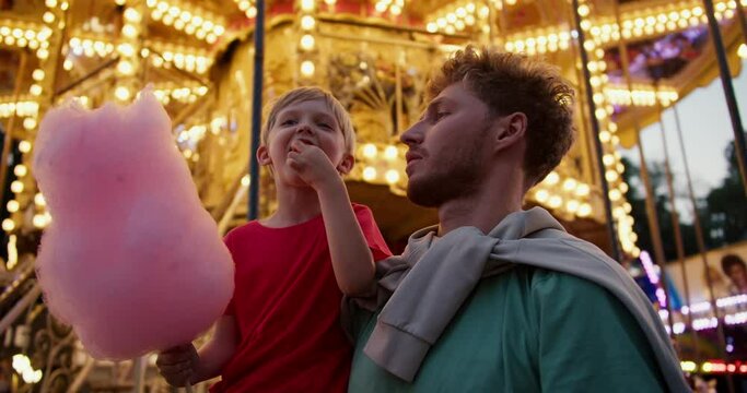 A little blond boy in a red T-shirt eats pink cotton candy with his dad in a Green T-shirt against the backdrop of a beautiful bright attraction with yellow flowers in an amusement park in the evening