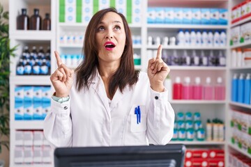 Middle age brunette woman working at pharmacy drugstore amazed and surprised looking up and pointing with fingers and raised arms.