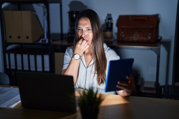 Young brunette woman working at the office at night smelling something stinky and disgusting, intolerable smell, holding breath with fingers on nose. bad smell