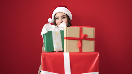 Beautiful hispanic woman wearing christmas hat holding gifts over isolated red background
