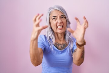 Fototapeta na wymiar Middle age woman with tattoos standing over pink background shouting frustrated with rage, hands trying to strangle, yelling mad