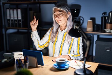 Middle age woman with grey hair working at the office at night with a big smile on face, pointing with hand and finger to the side looking at the camera.
