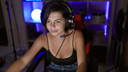 Confident young hispanic woman, beautiful streamer, engrossed in game, streaming in dark gaming room, technology glowing, wearing headphones, ebullient smile lighting up the night