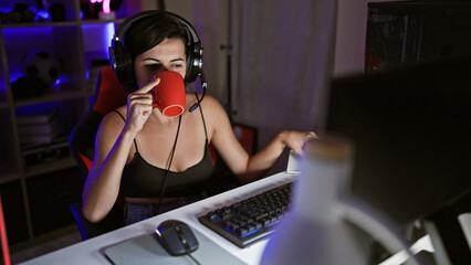 Radiant young hispanic woman streamer, in her cosy gaming room, playing a futuristic video game, sipping coffee amidst the ambient digital entertainment