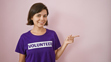 Charming young hispanic woman volunteer gleefully points to the side, presenting over isolated pink background