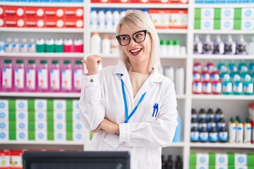 Young caucasian woman working at pharmacy drugstore smiling with happy face looking and pointing to the side with thumb up.