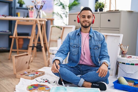 Young hispanic man painter sitting on the floor at art studio looking positive and happy standing and smiling with a confident smile showing teeth