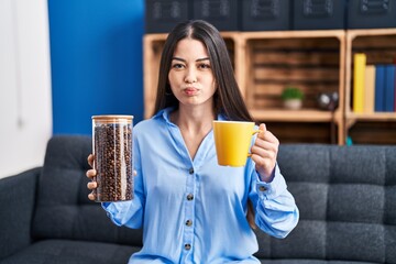 Young brunette woman holding coffee beans and cup of coffee puffing cheeks with funny face. mouth inflated with air, catching air. - obrazy, fototapety, plakaty