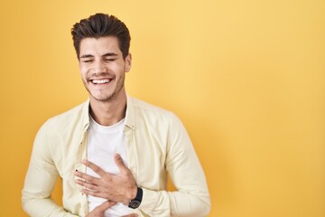 Young hispanic man standing over yellow background smiling and laughing hard out loud because funny crazy joke with hands on body.