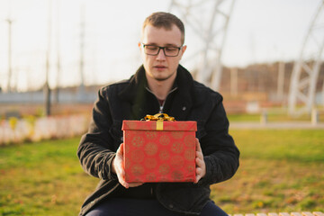 a young handsome man wearing eyeglasses giving red gift box to girlfriend outdoors