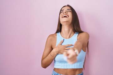 Young brunette woman standing over pink background laughing at you, pointing finger to the camera...