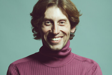 Male beauty concept. Emotional portrait of laughing handsome man in violet knitted polo neck posing over light blue background. Glossy long hair, shiny perfect smile. Studio shot
