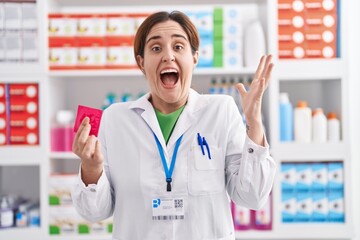 Brunette woman working at pharmacy drugstore holding condom celebrating victory with happy smile...