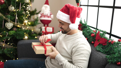 Obraz na płótnie Canvas Young hispanic man unpacking gift sitting on sofa by christmas tree at home