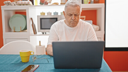 Middle age grey-haired man using laptop with relaxed expression at dinning room