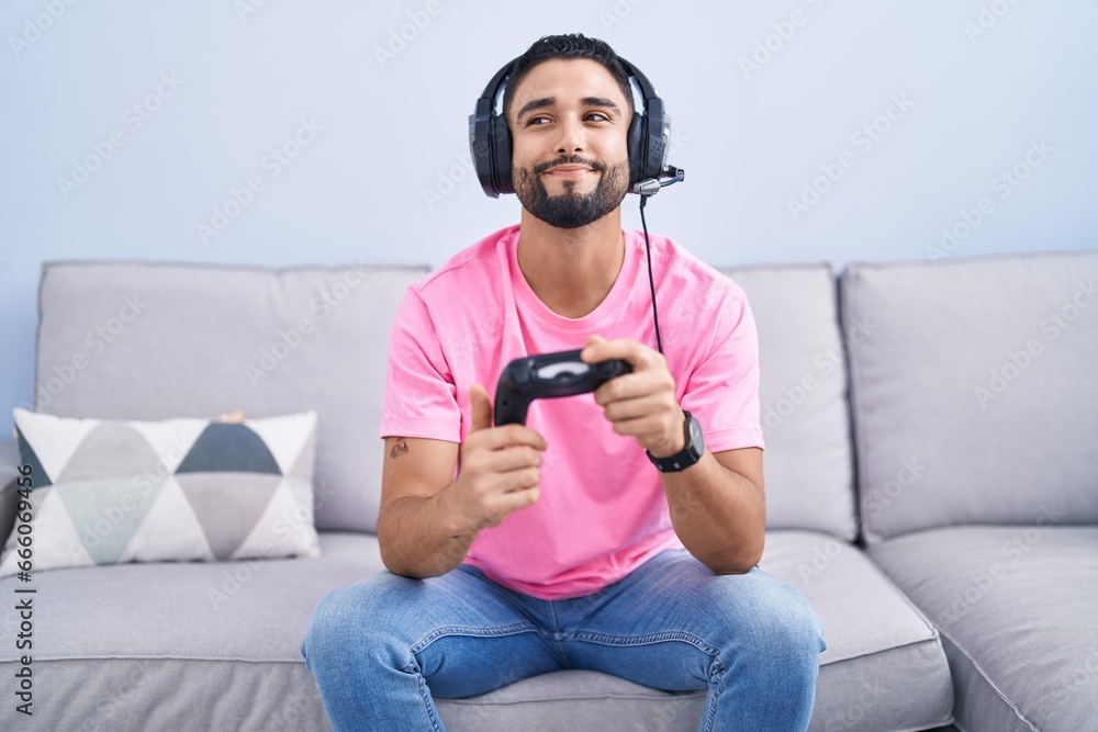 Poster Hispanic young man playing video game holding controller sitting on the sofa smiling looking to the side and staring away thinking.