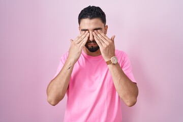 Hispanic young man standing over pink background rubbing eyes for fatigue and headache, sleepy and...