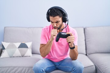 Hispanic young man playing video game holding controller sitting on the sofa feeling unwell and coughing as symptom for cold or bronchitis. health care concept.