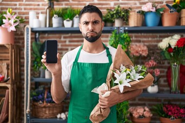 Hispanic young man working at florist shop showing smartphone screen puffing cheeks with funny face. mouth inflated with air, catching air. - obrazy, fototapety, plakaty