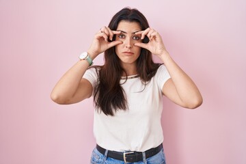 Young brunette woman standing over pink background trying to open eyes with fingers, sleepy and tired for morning fatigue