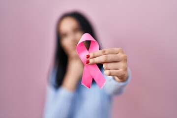 Hispanic woman holding pink cancer ribbon serious face thinking about question with hand on chin,...
