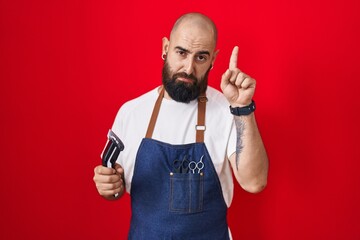 Young hispanic man with beard and tattoos wearing barber apron holding razor pointing up looking...