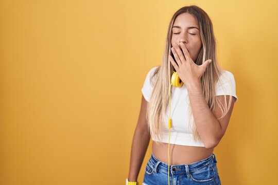 Young blonde woman standing over yellow background wearing headphones bored yawning tired covering mouth with hand. restless and sleepiness.