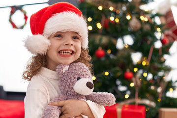 Fototapeta na wymiar Adorable blonde girl hugging teddy bear standing by christmas tree at home