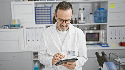 Middle age man with grey hair, a dedicated scientist, engrossed in his research on a touchpad at the heart of a busy lab