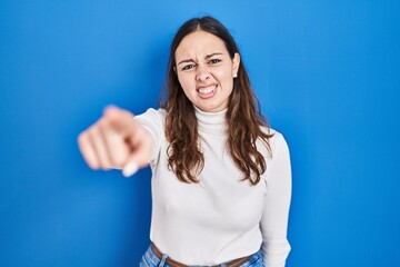 Young hispanic woman standing over blue background pointing displeased and frustrated to the...