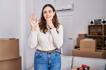 Young hispanic woman holding keys of new home serious face thinking about question with hand on...
